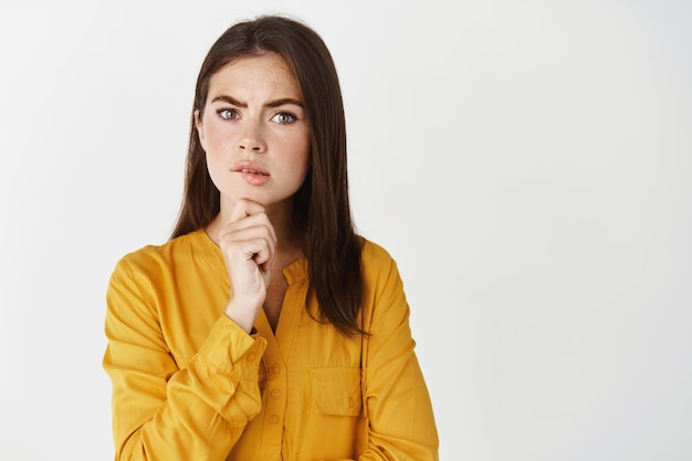 Close-up of serious young woman having doubts, looking at front thoughtful and doubtful, standing over white wall