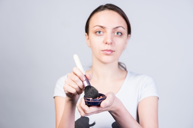 Chiuda sulla giovane donna seria che tiene un trucco fondotinta in polvere e un pennello mentre guarda la telecamera.