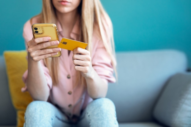 Close-up of serious young woman paying something online with her credit card and the smartphone while sitting on sofa at home.