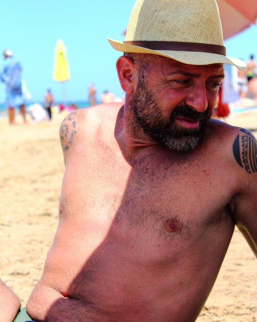 Close-up of shirtless man wearing sun hat while sitting at beach on sunny day