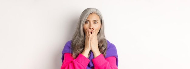 Close up of shocked asian old woman with grey hair standing in stylish pink sweater gasping startled