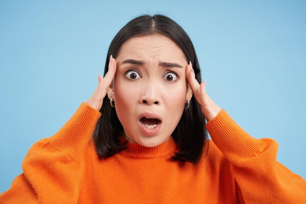 Close up of shocked asian woman in panic anxious looks terrified stands over blue studio background