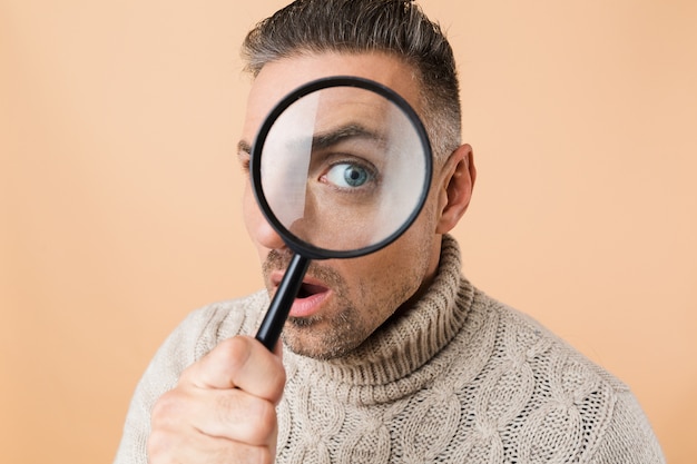 Close up of a shocked man looking through a magnifying glass isolated over beige wall