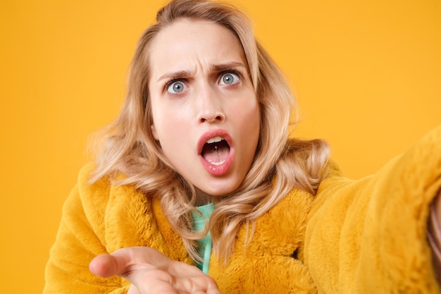 Close up of shocked perplexed young blonde woman in yellow fur coat posing isolated on orange background in studio. people lifestyle concept. mock up copy space. doing selfie shot on mobile phone