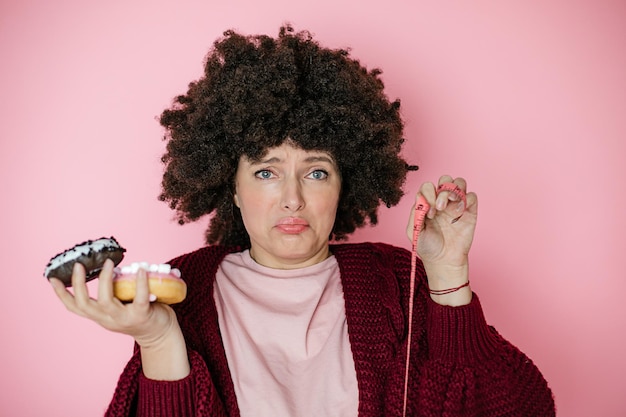 Close-up of shocked and upset beautiful afro hairstyle woman holding donut in hands and measuring tape. Concept of rejection of high-calorie foods and healthy lifestyle choice. addiction concept