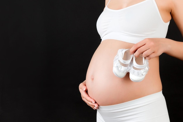 Close up of shoes for baby girl in pregnant womans hands at black background Future mother is wearing white underwear Motherhood concept Copy space