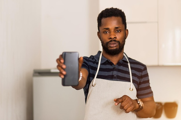 Immagine ravvicinata di attraente barbuto giovane afroamericano in grembiule che mostra smartphone con schermo nero vuoto in piedi in cucina. messa a fuoco selettiva. cucina, tecnologia, concetto pubblicitario.