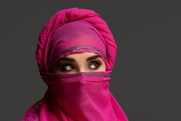 Close-up shot of a beautiful girl with an expressive smoky eyes wearing an elegant pink hijab decorated with sequins. She is posing at the studio and looking away on a dark background. Human emotions,