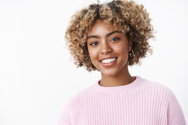 Photo close-up shot of charming good-looking stylish african-american woman with piercing and blond curly haircut smiling delighted and friendly at camera wearing warm sweater posing joyful over white wall