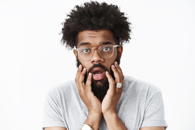 Close-up shot of concerned and worried adult african american bearded guy with afro hairstyle open mouth shook and touching face as being frustrated and troubled over gray wall