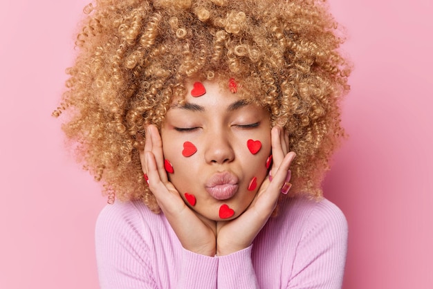 Close up shot of curly haired romantic young woman keeps lips\
rounded sends mwah keeps eyes closed wears casual jumper isolated\
over pink background wants to kiss her boyfriend on valentines\
day