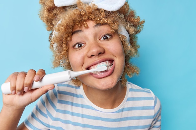 Close up shot of curly haired woman brushes teeth with toothpaste for having fresh breath looks with glad expression at camera undergoes whitening procedures at home poses against blue wall