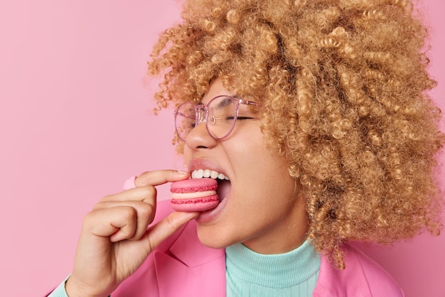 Close up shot of curly woman bites macaroon hungrily enjoys eating sweet dessert keeps mouth opened wears spectacles and formal outfit isolated over pink background Unhealthy food eating extra sugar