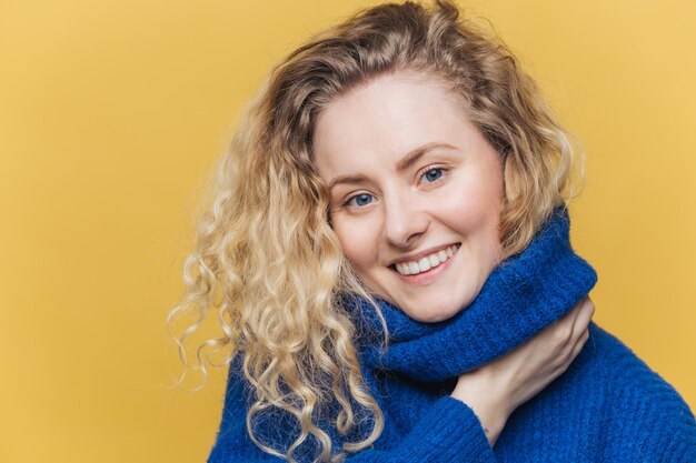 Close up shot of delighted satisfied female model has curly blonde hair broad smile demonstrates white perfect teeth wears blue knitted sweater isolated over yellow background Happiness