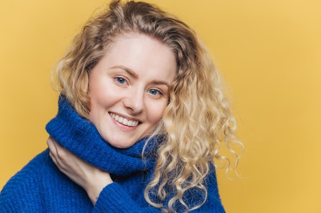 Close up shot of delighted satisfied female model has curly\
blonde hair broad smile demonstrates white perfect teeth wears blue\
knitted sweater isolated over yellow background happiness