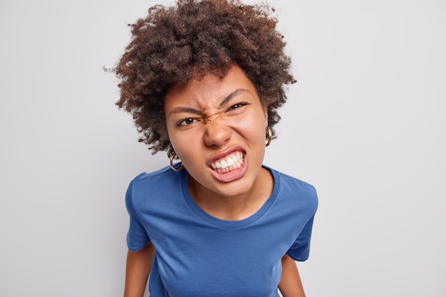 Close up shot of displeased African American woman frowns face clenches teeth from anger dressed in casual blue t shirt expresses negative emotions poses against white background. Anger concept