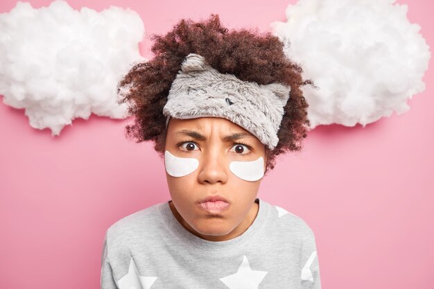 Close up shot of displeased woman with curly hair stares shocked at camera wears blindfold and pajama isolated over pink wall has bad mood after awakening