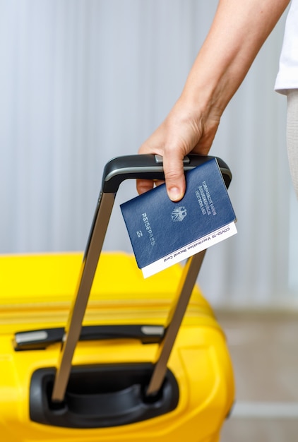 Close up shot of female hand holding deutschland passport coronavirus covid-19 vaccination record card certificate and pull yellow trolley luggage bag to traveling abroad after lockdown quarantine