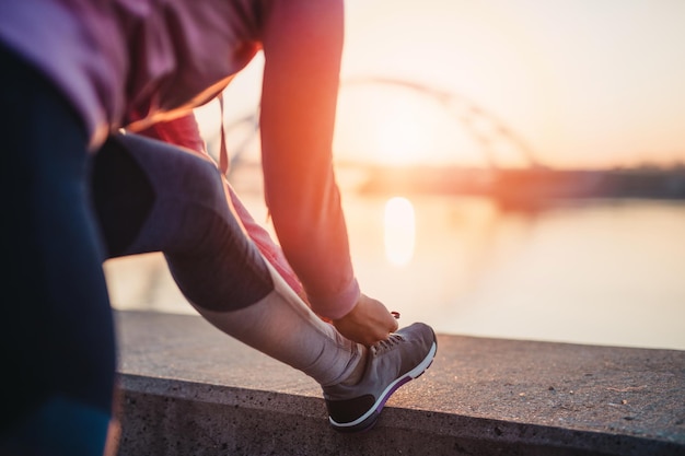 スニーカーで靴を結ぶ女性のジョガーのクローズアップショット。美しい川の橋と背景の夕日。