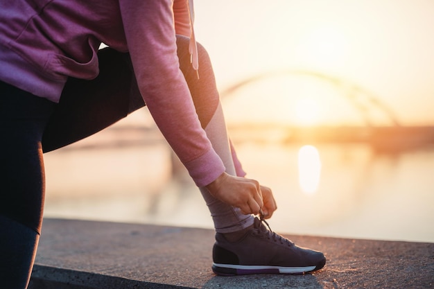 Close up shot of female jogger ties shoes on sneakers. Beautiful river bridge and sunset in the background.