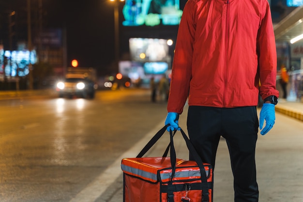 Close-up shot of a food deliveryman in red uniform carrying a food delivery box to deliver for customer for order during COVID-19 outbreak lockdown in the city at night time