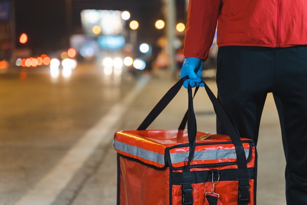 Close-up shot of a food deliveryman in red uniform carrying a food delivery box to deliver for customer for order during COVID-19 outbreak lockdown in the city at night time