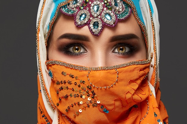 Close-up shot of a gorgeous girl with professional make-up wearing an elegant colorful hijab decorated with sequins and jewelry. She is posing at the studio and looking at the camera on a dark backgro
