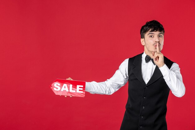 Close up shot of guy waiter in a uniform with butterfly on neck showing sale icon making silence gesture on isolated red surface