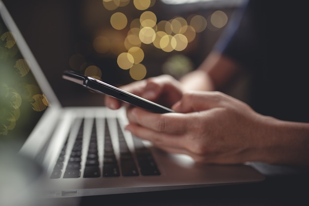 Close up shot of hands using smart phone typing message for searching with laptop