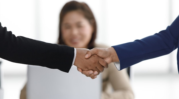 Close up shot of handshaking of unrecognizable unidentified successful businessman and businesswoman in formal suit in front female officer staff working with laptop computer in blurred background.