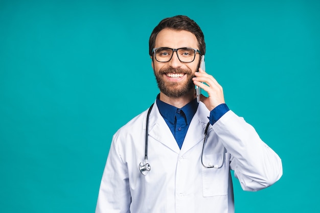 Close up shot of handsome young man doctor isolated on blue background talking on smartphone, smiling positively. Using mobile phone.