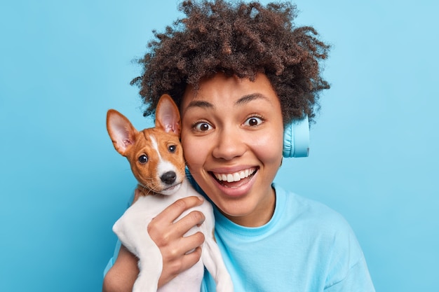 Close up shot of happy Afro American woman holds lovely pet near face glad to get pedigree dog as present on birthday have friendly relationships listens music in headphones isolated on blue