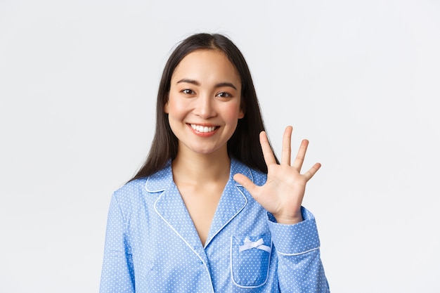 Close-up shot of happy attractive asian woman in blue pyjama showing five fingers and smiling white teeth, explain main rules or making order, standing white background delighted