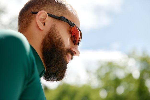 Close up shot of happy attractive young european man with beard\
wearing glasses and green sportswear looking aside during evening\
ride in park on weekend