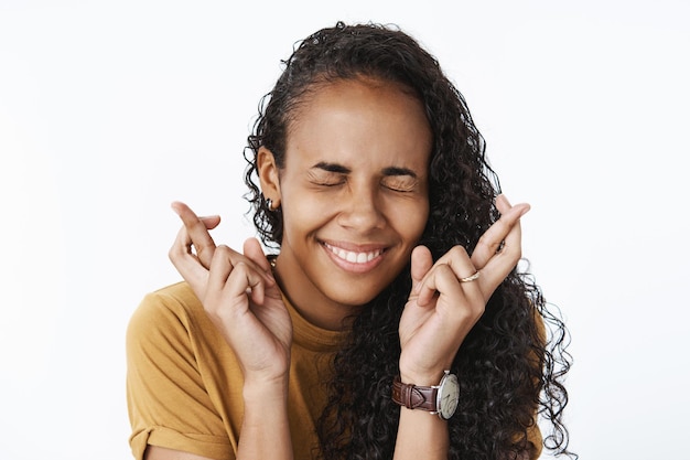 Close-up shot of happy excited and optimistic young african-american woman making wish with smile and closed eyes crossing fingers for good luck as anticipating dream come true over gray wall.