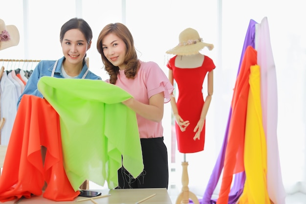 Закройте снимок счастливых двух красивых азиатских модельеров женского пола, выбирающих ткань из разнообразной ткани для шитья своей новой коллекции вместе.