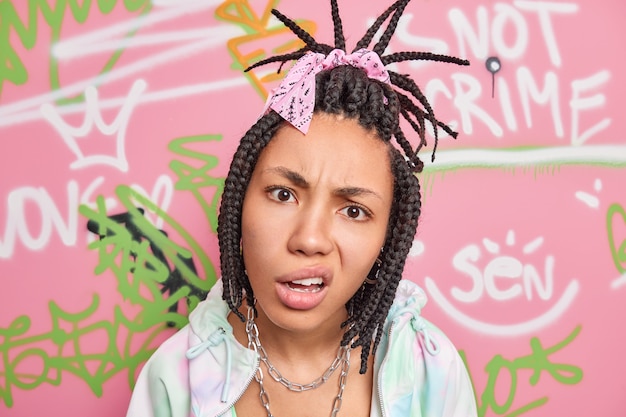 Close up shot of indignant grumpy hipster girl with combed dreadlocks smirks face dressed in fashionable clothes poses against graffiti wall poses in urban place