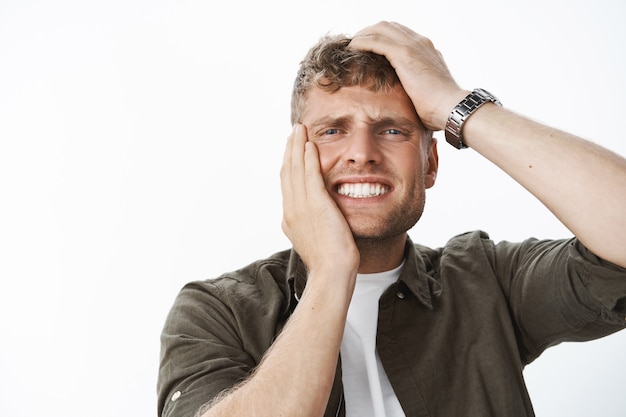 Close-up shot of man in agony and sorrow, feeling dismay and concern holding hands on head and face clenching teeth from painful feelings of regret and disappointment grimacing over gray wall