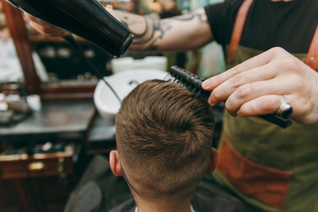 Immagine ravvicinata dell'uomo che ottiene taglio di capelli alla moda al negozio di barbiere. l'hairstylist maschile in tatuaggi che serve cliente, asciugando i capelli con un asciugacapelli