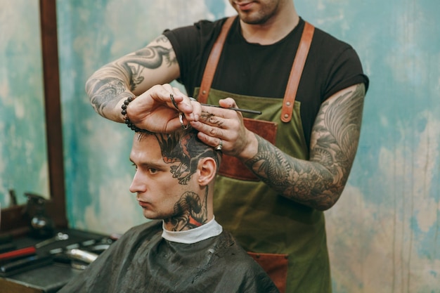 이발소에서 트렌디한 머리를 자르는 남자의 사진을 클로즈업하세요. 고객에게 봉사하는 문신의 남성 미용사.