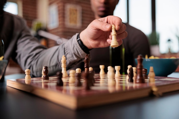 Close up shot of man hand moving chess piece on chessboard while sitting at table. smart person playing strategy boardgames with friends while sitting at home in living room.