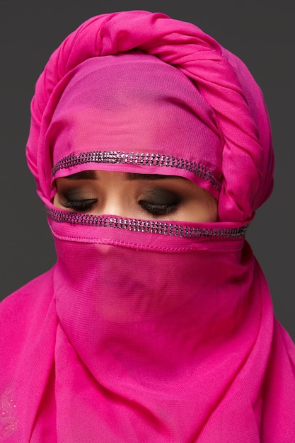 Close-up shot of a nice young female with an expressive smoky eyes wearing an elegant pink hijab decorated with sequins. She has turned her head and closed her eyes a dark background. Human emotions,