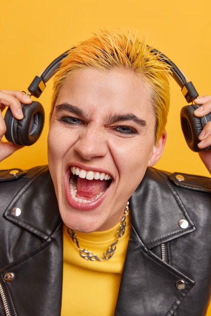 Close up shot of overjoyed female teenager with trendy hairstyle vivid makeup takes off headphones with high volume wears black leather jacket has sassy expression isolated over yellow wall