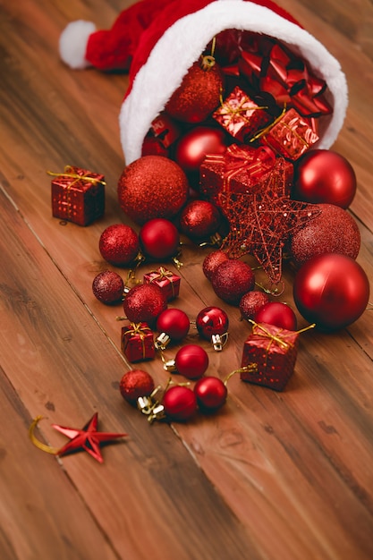 Close up shot of red decorative hanging items star shiny glossy sphere balls ribbon bow tie and present gift boxes pouring from Santa claus Christmas hat on dark brown wooden table with copy space.