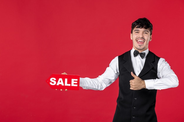 Close up shot of smiling guy waiter in a uniform with butterfly on neck showing sale icon making ok gesture on isolated red surface
