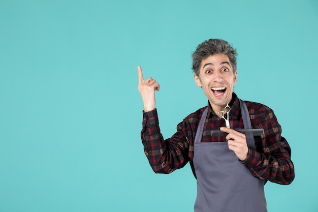 Close up shot of smiling young barber wearing gray apron and holding comb and scissor pointing up on blue color background