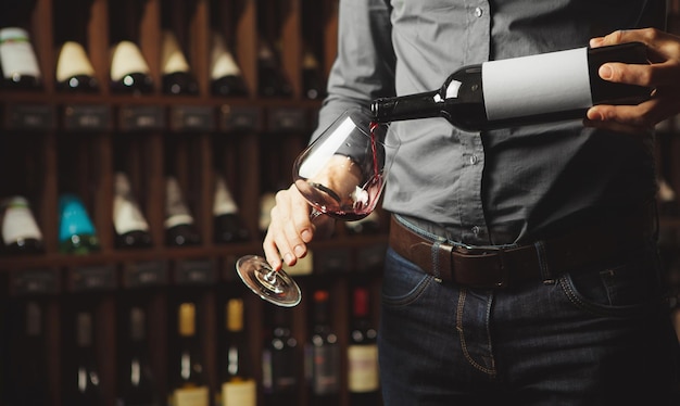 Close up shot of sommelier pouring red wine from bottle in glass on underground cellar background