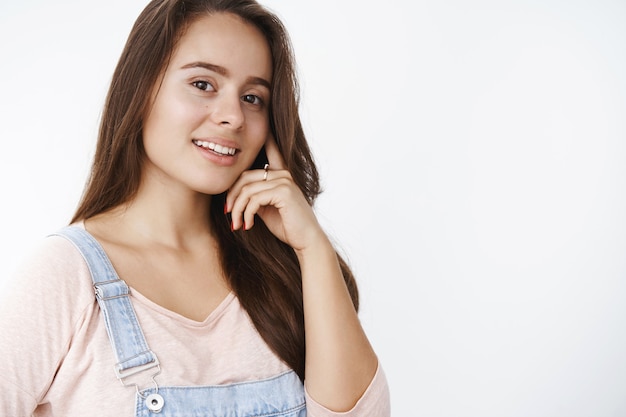 Close-up shot of stylish confident and tender woman with pure clear skin and brown hair touching cheek softly smiling caring at front posing delighted and sensual over gray wall