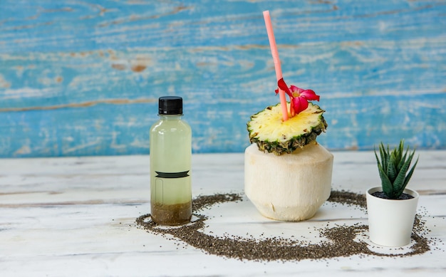 Close up shot of summer tropical coconut juice decorated with sliced sweet and sour pineapple blossom flower and straw placed on white and blue wooden table with bottle packaging for advertisement.