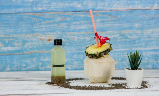 Close up shot of summer tropical coconut juice decorated with sliced sweet and sour pineapple blossom flower and straw placed on white and blue wooden table with bottle packaging for advertisement.